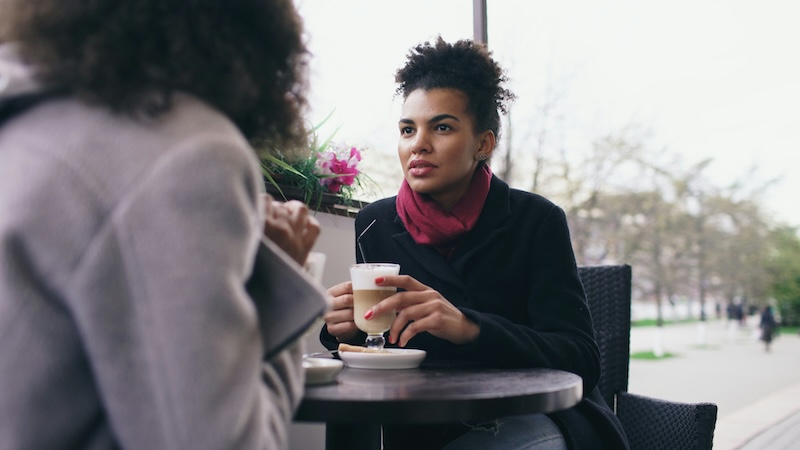 Two coworkers talk over coffee at an outdoor cafe table, the kind of open door policy conversation that happens when an employee feels safe speaking up.